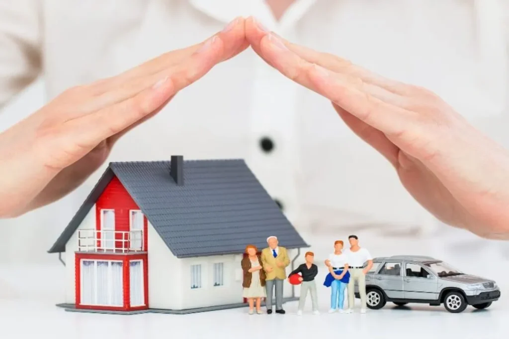 Mãos em formato de telhado protegendo uma maquete de casa, um carro em miniatura e uma família, simbolizando a segurança patrimonial e a quitação do consórcio através do seguro prestamista em caso de imprevistos.